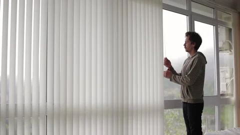 Man adjusting vertical window blinds, filtering daylight streaming through Stock Footage 308851774