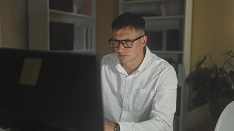 Man adjusts glasses while working at a computer in an office with shelves a.. Stock Footage 347714672