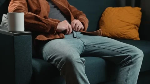 A man adjusts his clothing while sitting on a couch with cushions. The setting Stock-Footage 339097571