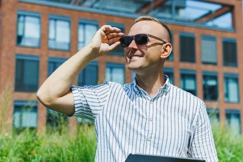 Man adjusts sunglasses while standing outside a building on a sunny day Stock Photos