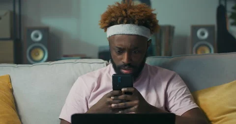 Man of African appearance is sitting on sofa, holding phone and laptop. He 스톡 동영상 218115653