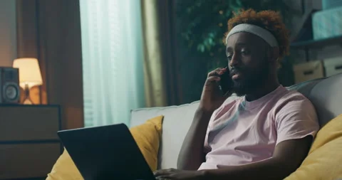 Man of African appearance is sitting on sofa, holding laptop on lap and writing Stock Footage 218122261