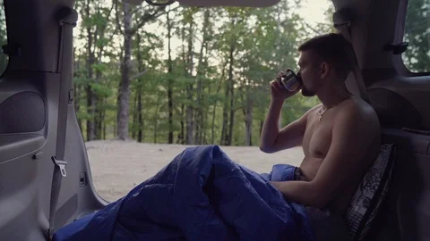 A man after sleeping sits in the trunk of a minivan and drinks hot tea and Stock Footage 117628155