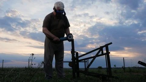 A man in age cuts off a metal angle grinder at sunset. Beautiful sky behind the Stock Footage 76917169