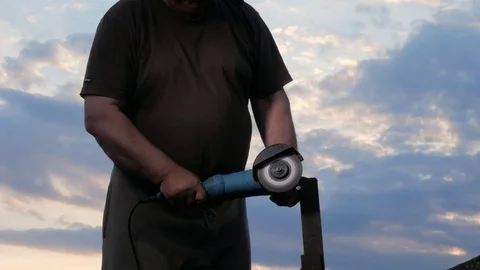 A man in age cuts off a metal angle grinder at sunset. Beautiful sky behind the Stock Footage 76917318