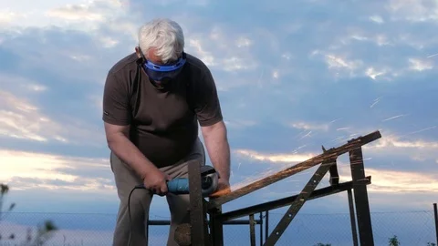 A man in age cuts off a metal angle grinder at sunset. Beautiful sky behind the Stock Footage 76917443