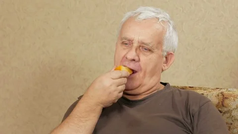 Man aged overweight eats fruit at home on the couch. He cut off a piece of Stock Footage 71244657