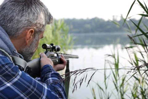 Man aiming with hunting rifle near lake outdoors. Space for text Фото