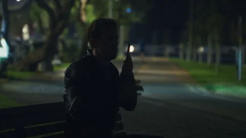 Man alone on a park bench at night, speaking on his phone while scanning the 스톡 동영상 311616531