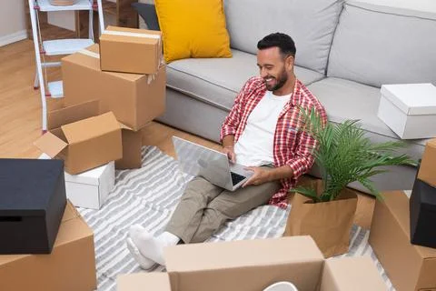 Man, amidst moving boxes, uses a laptop to shop for decor, envisioning his new Stock Photos