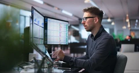 Man Analyzes Medical Billing Data On Dual Laptop Screens In Busy Office. Stock Photos