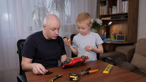 A man and a boy are sitting together at a table and repairing a broken toy car. Stock Footage 327644549