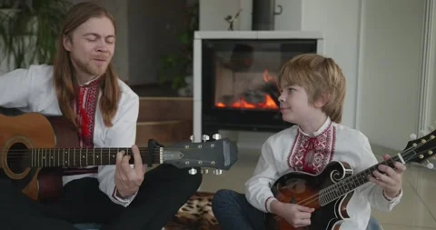 Man and Kid Boy Play Music on Guitar and Mandolin Sitting on Floor by Fireplace Stock Footage 337227321