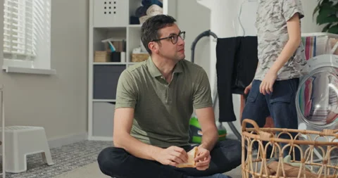 Man and son sit on laundry room floor relaxing after sorting laundry, eating 库存影片 200844332