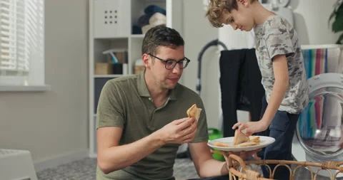Man and son sit on laundry room floor relaxing after sorting laundry, eating Видео 200844955