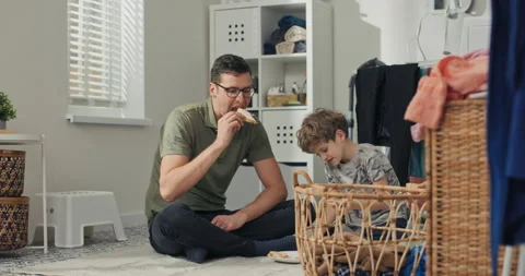 A man and son sit on the laundry room floor relaxing after sorting laundry 库存影片 200845817