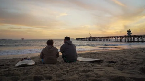 Man and son sitting on beach with surfboard at sunset Stock Footage 105621694