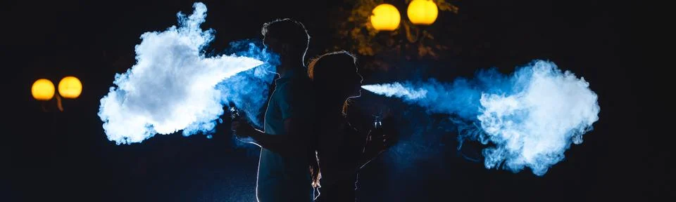 The man and woman smoke an electrical cigarette on the dark street. night tim Stock Photos