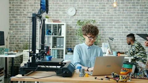 Man and woman using tablet, laptop and 3d printer in office talking working Stock Footage 119765358