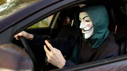 A man in an anonymous mask sits behind the wheel of a car and uses a smartphone Stockbeeldmateriaal 163777752