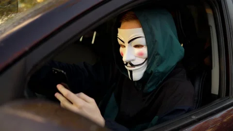 A man in an anonymous mask sits behind the wheel of a car and uses a smartphone Stock Footage 163778541