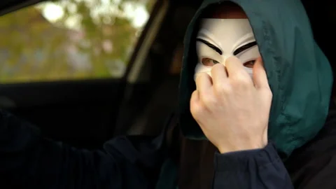 A man in an anonymous mask sits at the wheel of a car and takes off his Stock Footage 163778014