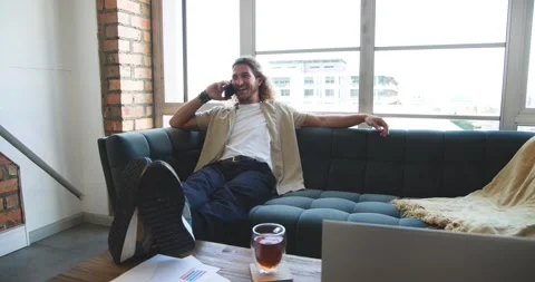 Man answering incoming work call, reclining on dark teal sofa in bright loft Stock Footage 339959351