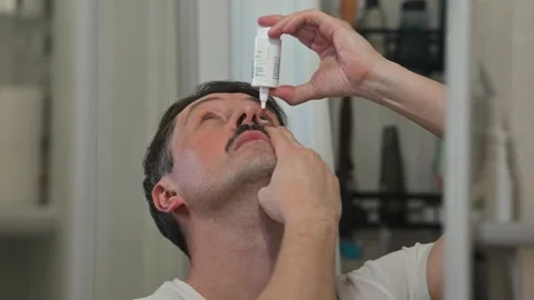 A man applies eye drops while looking in a mirror. The setting features a 스톡 동영상 337111672