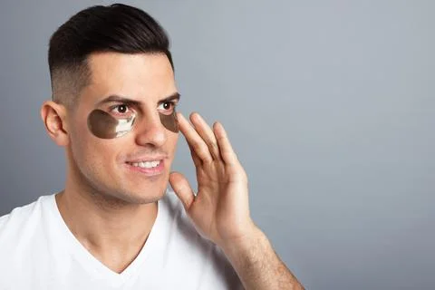 Man applying dark under eye patch on grey background. Space for text Stock Photos