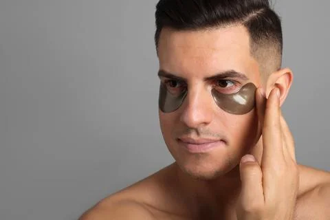 Man applying dark under eye patch on grey background. Space for text Stock Photos
