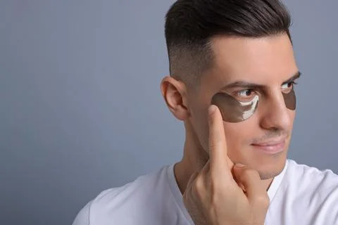 Man applying dark under eye patch on grey background. Space for text Stock Photos