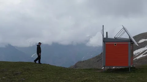 Man approaches modern cabin in mountainous landscape with cloudy sky backdrop Stock Footage 336964816