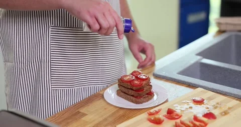 Man in apron pours salt from jar on stack of bread and tomato slices 스톡 동영상 284489607