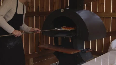 A man in an apron is putting raw pizza into a wood-fired pizza oven. Cooking Stock Footage 259063196