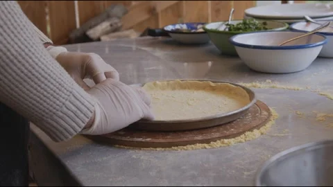 A man in an apron is rolling out pizza dough on a table sprinkled with flour. On Stock Footage 259405706