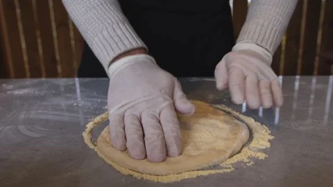 A man in an apron is rolling out pizza dough on a table sprinkled with flour. On Stock Footage 259695989