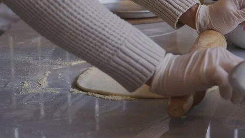 A man in an apron is rolling out pizza dough on a table sprinkled with flour. On Stock Footage 259700342