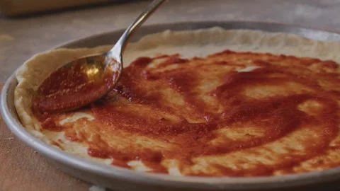 A man in an apron is spreading tomato paste on a pizza dough base on a table Stock Footage 258649627