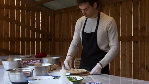 A man in an apron is spreading tomato paste on a pizza dough base on a table Stock Footage 258844960