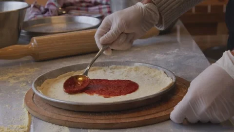 A man in an apron is spreading tomato paste on a pizza dough base on a table Stock Footage 259063720