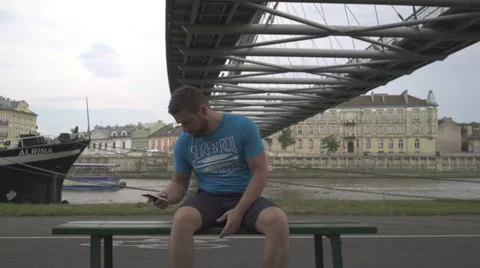 Man arguing on the cellphone, sitting on the bench against the river. 库存影片 65187798