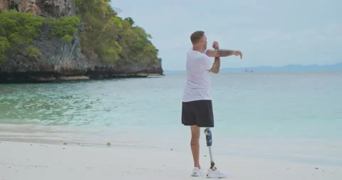 Man with artificial bionic leg doing body warm up before workout on a beach. Video stock 242464115