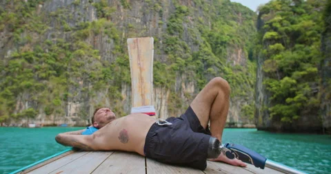 Man with artificial bionic leg relaxing by laying down on long tail boat front. Video stock 201366569