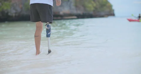 Man with artificial bionic legs standing on beach kicks up water in slow motion. Video stock 195000242