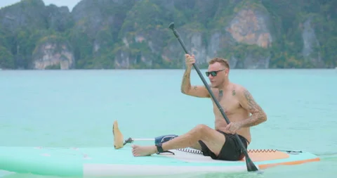 Man with artificial leg sitting on paddle board and sailing at tropical sea. Video stock 187699530