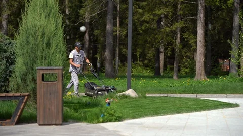A man of Asian appearance in a gray work suit and a black cap mows the grass Stock Footage 108985457