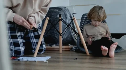 A man assembles a chair while his little son sits nearby and watches a tablet Stock Footage 305946567