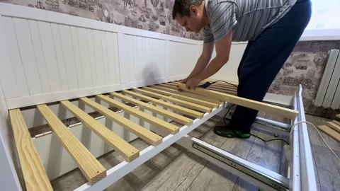 A man is assembling an ottoman at home. He attaches wooden slats to the bas.. Video stock 304378284