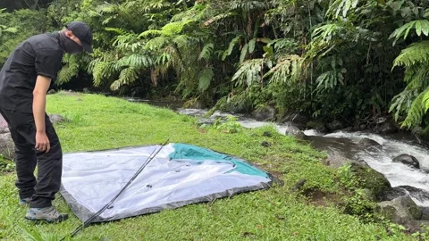 Man Assembling Tent Poles by Rapid River in Forest, Static Shot Stock Footage 323676110