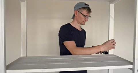 Man Assembling White Wooden Shelf Unit, Using Electric Screwdriver to Secure Stock Footage 314791582
